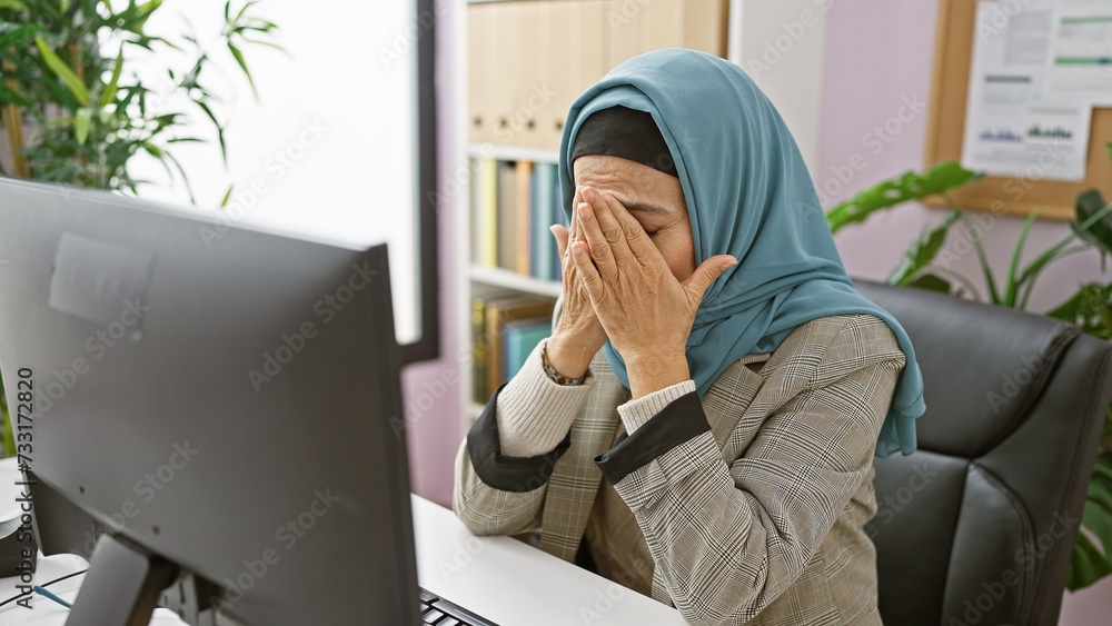 A stressed middle-aged woman in hijab feeling overwhelmed at her office workstation, eyes covered, expressing a moment of frustration or headache. A stressed middle-aged woman in hijab feeling overwhelmed at her office workstation, eyes covered, expressing a moment of frustration or headache.