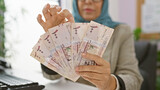 A woman in a hijab counts saudi riyal bills at her office, suggesting a business transaction or financial management indoors.
