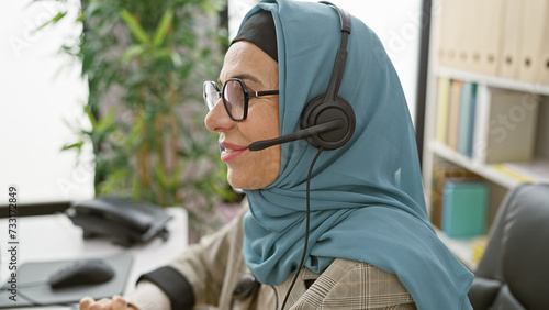 A professional woman wearing a hijab and headset working in a modern office environment.
