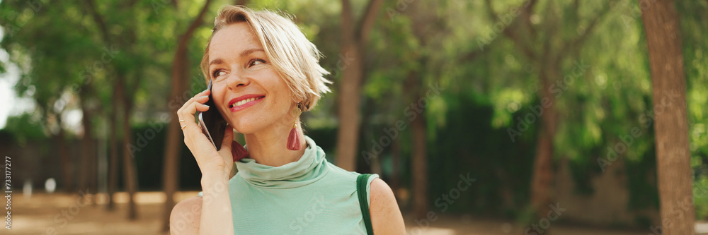 Panorama of beautiful smiling woman with short blond hair in casual clothes walks through the city square, talking on cellphone