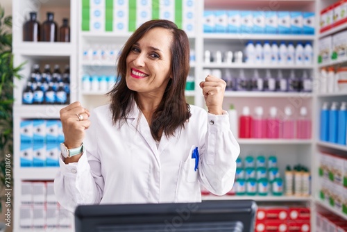 Middle age brunette woman working at pharmacy drugstore very happy and excited doing winner gesture with arms raised, smiling and screaming for success. celebration concept.