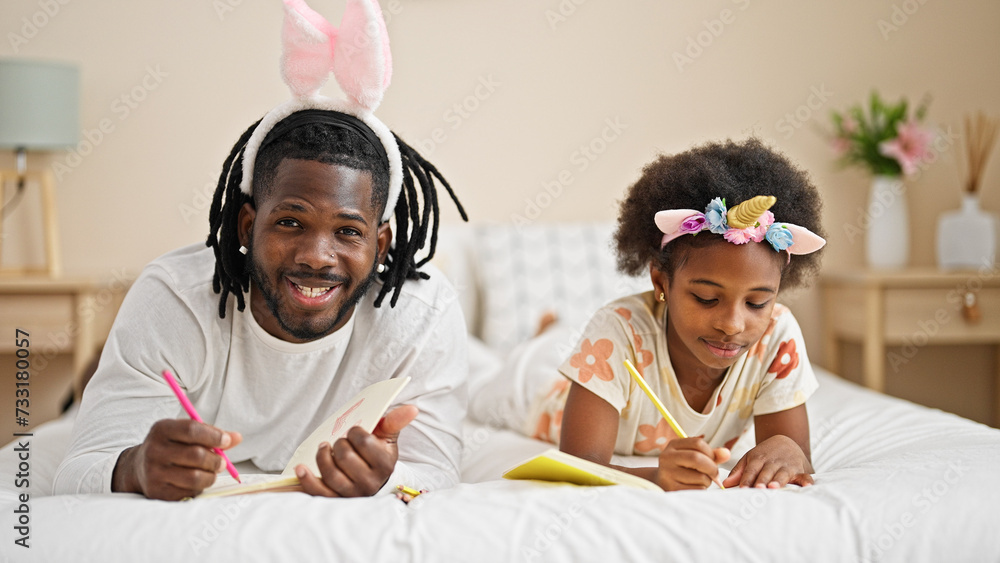 African american father and daughter wearing funny diadem drawing on notebook lying on bed at bedroom - obrazy, fototapety, plakaty 