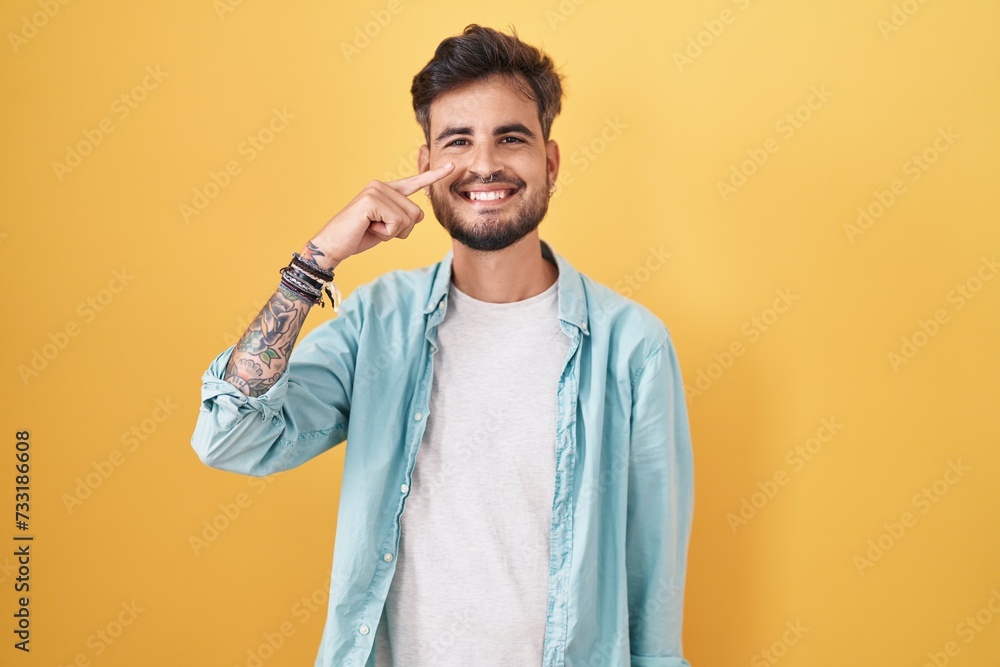 Young hispanic man with tattoos standing over yellow background pointing with hand finger to face and nose, smiling cheerful. beauty concept Young hispanic man with tattoos standing over yellow background pointing with hand finger to face and nose, smiling cheerful. beauty concept