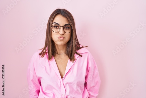 Young hispanic woman wearing glasses standing over pink background puffing cheeks with funny face. mouth inflated with air, crazy expression. photo
