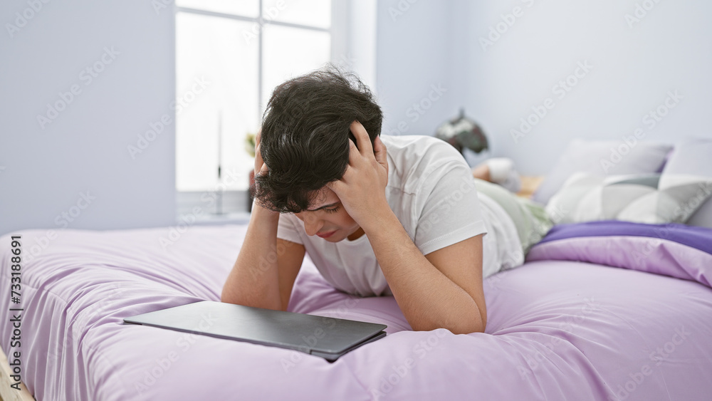 Distressed young man with laptop in a modern bedroom conveying frustration or headache. Distressed young man with laptop in a modern bedroom conveying frustration or headache.