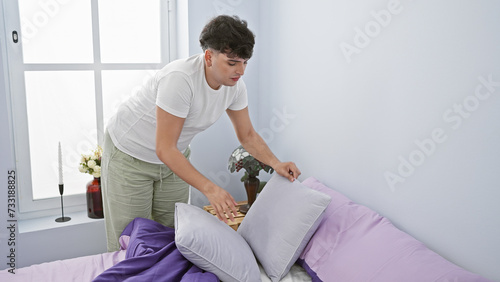 Young man organizing pillows on a bed in a bright, minimalist bedroom interior.