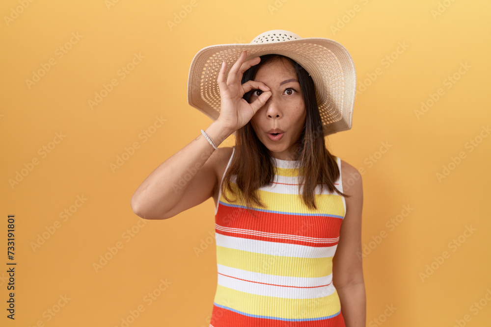 Middle age chinese woman wearing summer hat over yellow background doing ok gesture shocked with surprised face, eye looking through fingers. unbelieving expression. Middle age chinese woman wearing summer hat over yellow background doing ok gesture shocked with surprised face, eye looking through fingers. unbelieving expression.