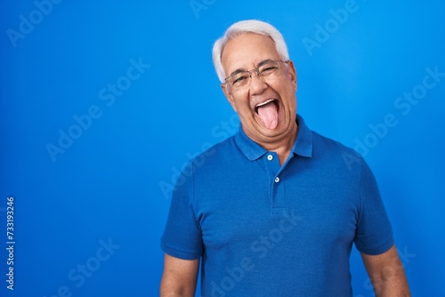 Middle age man with grey hair standing over blue background sticking tongue out happy with funny expression. emotion concept. © Krakenimages.com