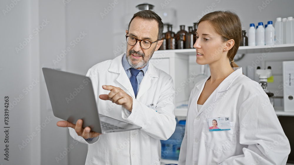 Two dedicated scientists working shoulder-to-shoulder, speaking over lab analysis on a laptop in the heart of a bustling medical research laboratory. Two dedicated scientists working shoulder-to-shoulder, speaking over lab analysis on a laptop in the heart of a bustling medical research laboratory.