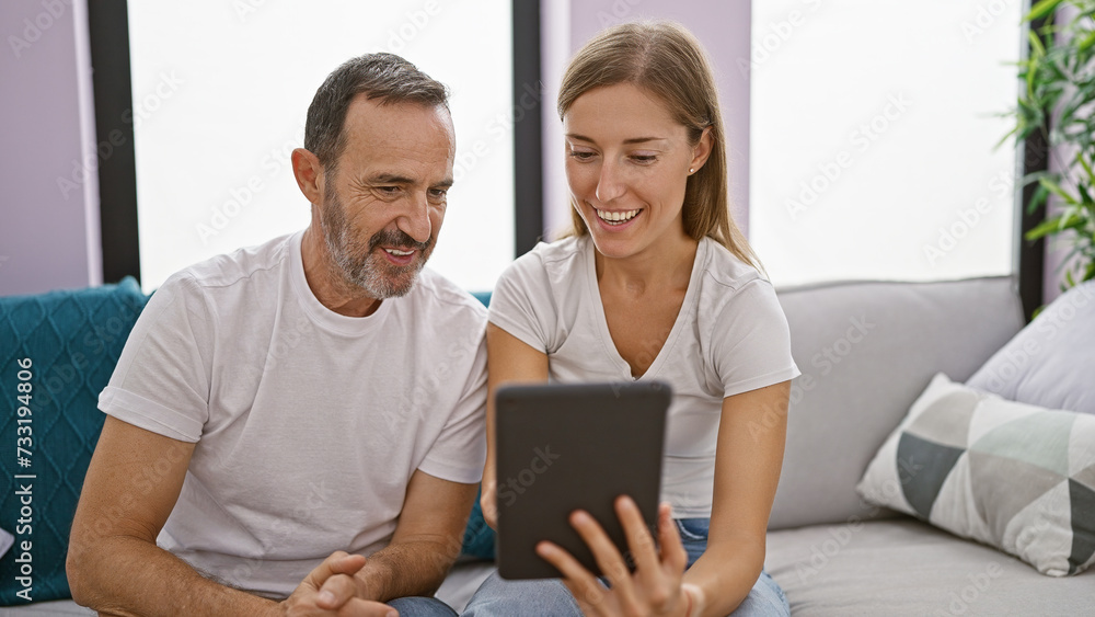 Father and daughter share a jovial moment, confidently smiling while sitting together on the homely sofa, fully engrossed in their touchpad, enjoying their relaxed online lifestyle. Father and daughter share a jovial moment, confidently smiling while sitting together on the homely sofa, fully engrossed in their touchpad, enjoying their relaxed online lifestyle.