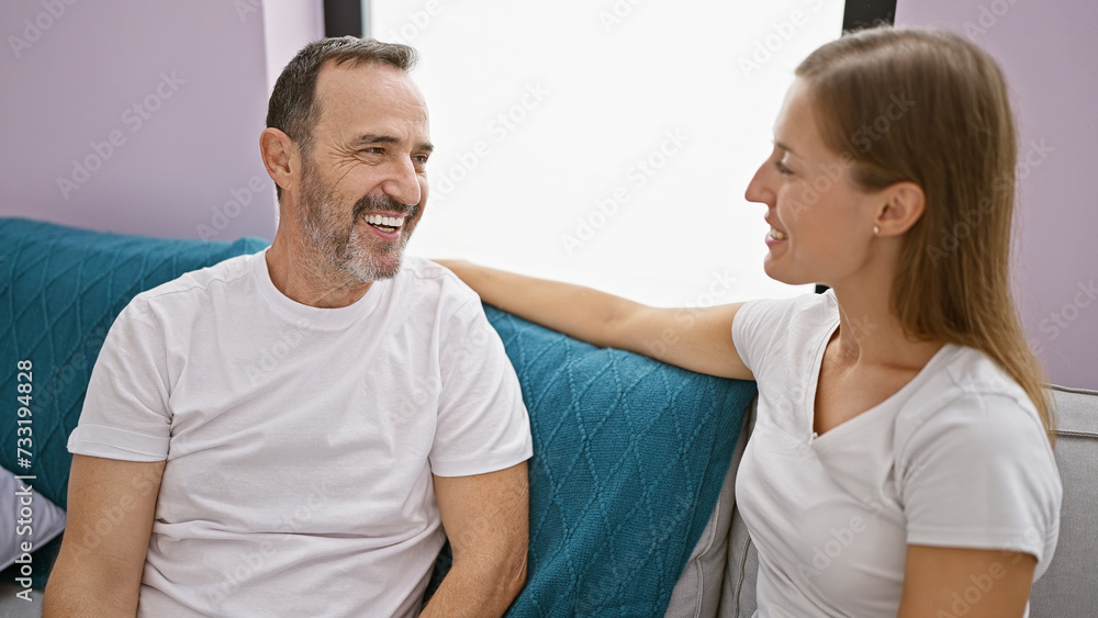 Fototapeta premium Confident father and joy-filled daughter sharing a fun, relaxed moment of love, sitting on the living room sofa at home, laughing and looking at each other, smiling in their casual lifestyle.