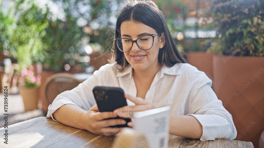 Fototapeta premium Young beautiful hispanic woman smiling happy using smartphone sitting on the table at bar terrace