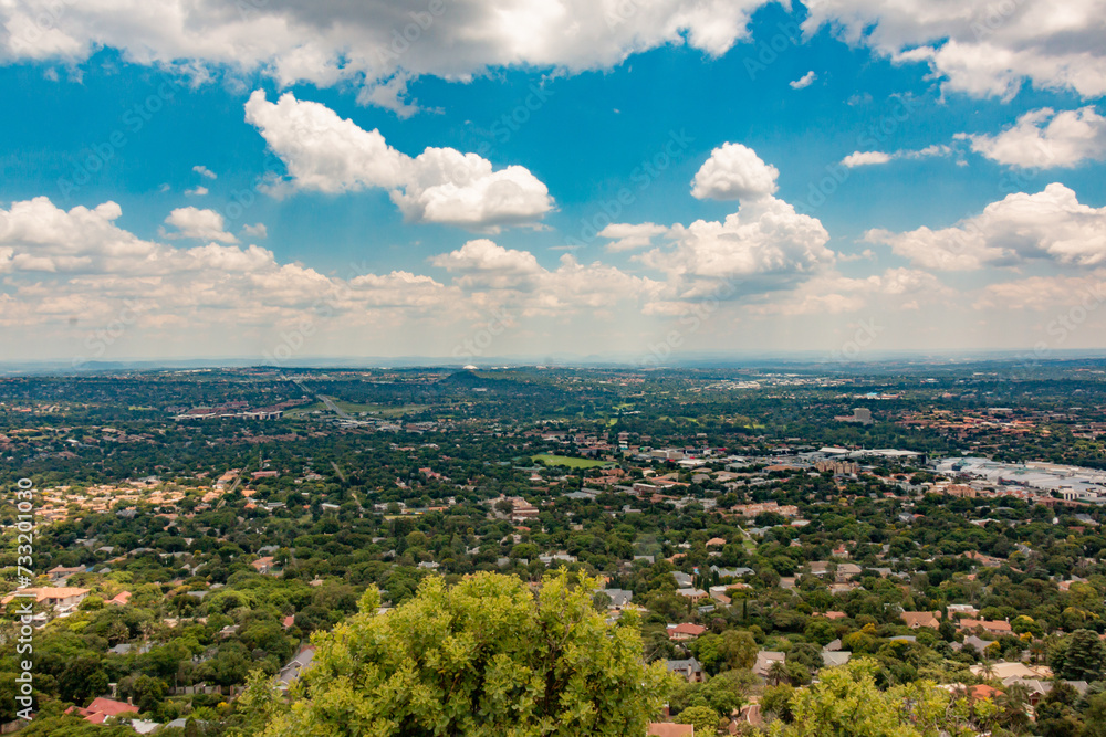 Fototapeta premium Clear Elevated Panoramic view of Johannesburg - Western View from Northcliff with trees in foreground and white clouds in a blue sky