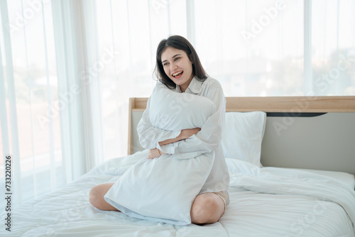 Photo of young happy woman in pajama stretching her arms and smiling while sitting on bed after sleep.