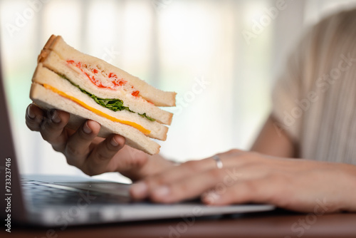 Close-up of a woman using a laptop while holding a tasty sandwich for the concept of fast food eating at the workplace