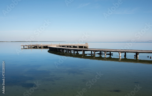 A wide footbridge made of wooden planks, from the side, on shallow water on the shore. The horizon with blue sky in the background.