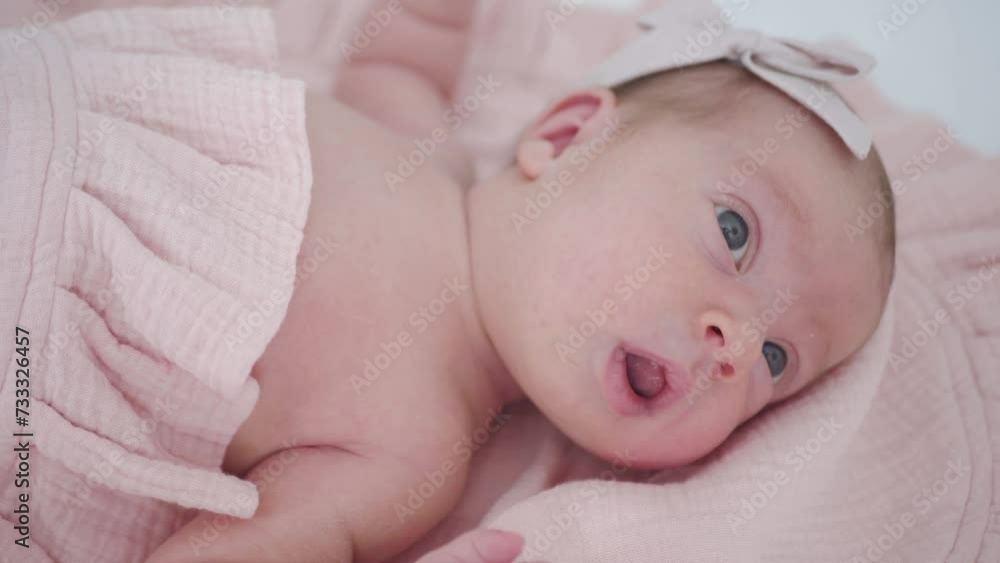 Portrait of a beautiful newborn baby girl lying in a white cot under a pink plaid with ruffles at home with a bow on her head and looking around. Portrait of a beautiful newborn baby girl lying in a white cot under a pink plaid with ruffles at home with a bow on her head and looking around.