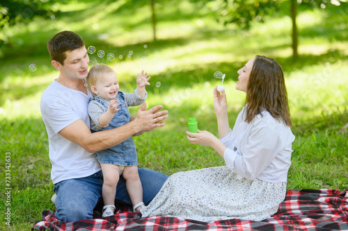 Happy family enjoying a picnic in the park in spring, blowing bubbles and smiling while sitting on a checkered blanket spread on the grass. Concept of a happy, playful, harmonious family relationship © GRON777