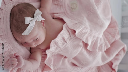 Portrait of a beautiful newborn baby girl lying in a white cot under a pink plaid with ruffles at home with a bow on her head and looking around. photo