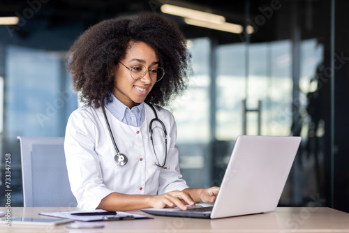 Confident african american female doctor in medical uniform working by laptop in workspace. Smiling therapist with stethoscope prescribing medication after web consultation of client in clinic. photo