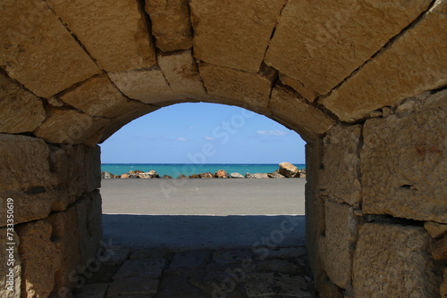 Arco de piedras a trav  s del cual se ve en el fondo una carretera asfaltada  piedras y el mar con el cielo.
