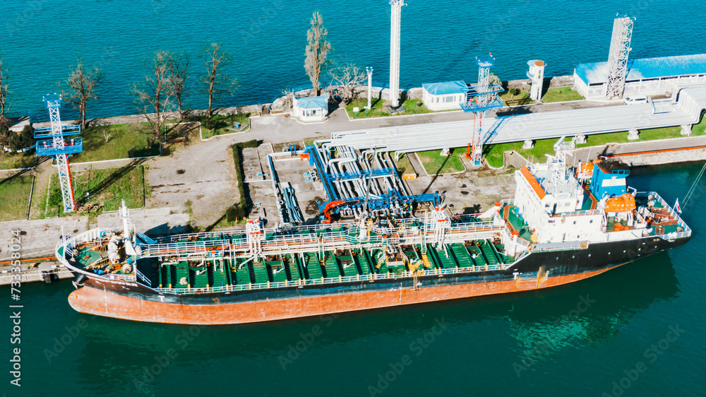 Aerial shot captures a large tanker ship moored at a port, surrounded by complex piping and fuel storage infrastructure. 