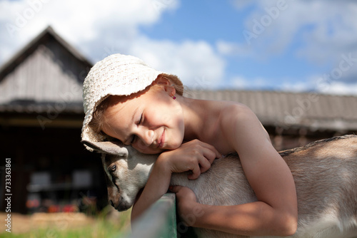 Happy girl hugs a baby goat. Hot summer day in the village. Heat, July, farm, farmstead.