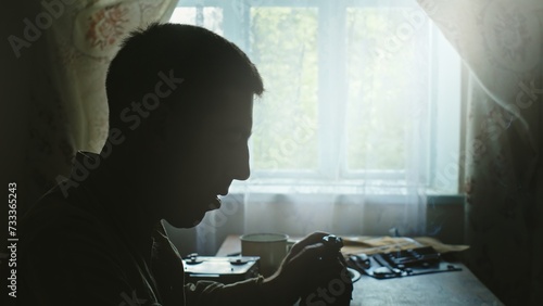 Soldier sitting drinking from an army water bottle at a table in a gloomy house facing a window with lace curtain