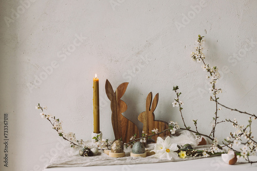 Happy Easter! Stylish easter eggs, wooden bunnies, cherry blossom and candle on rustic table. Modern natural dyed marble eggs and spring flowers. Easter still life decor in countryside home