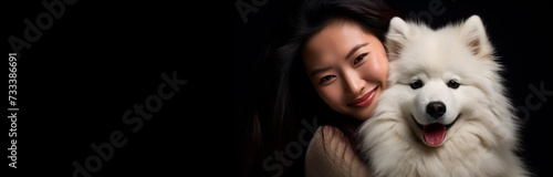 In the image, a woman with long dark hair is gently embracing a fluffy white dog, both appearing content with warm smiles against a black background. The woman's affectionate pose and the dog's relaxe