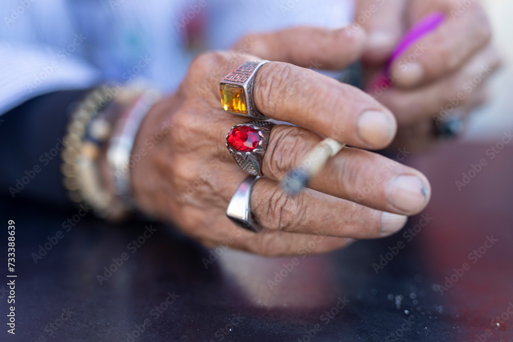 hands of an old man in a blue shirt with cheap jewelry, plastic stones and a cigarette on the table. Fragment. hands of an old man in a blue shirt with cheap jewelry, plastic stones and a cigarette on the table. Fragment.