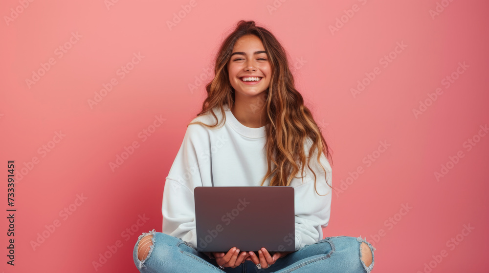 cheerful young woman sitting with her legs crossed, holding a laptop on her lap, wearing a white sweatshirt and ripped blue jeans, against a coral pink background cheerful young woman sitting with her legs crossed, holding a laptop on her lap, wearing a white sweatshirt and ripped blue jeans, against a coral pink background