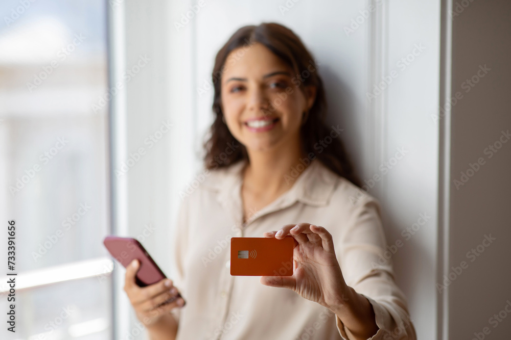 Confident young woman presenting credit card while holding smartphone at home Confident young woman presenting credit card while holding smartphone at home