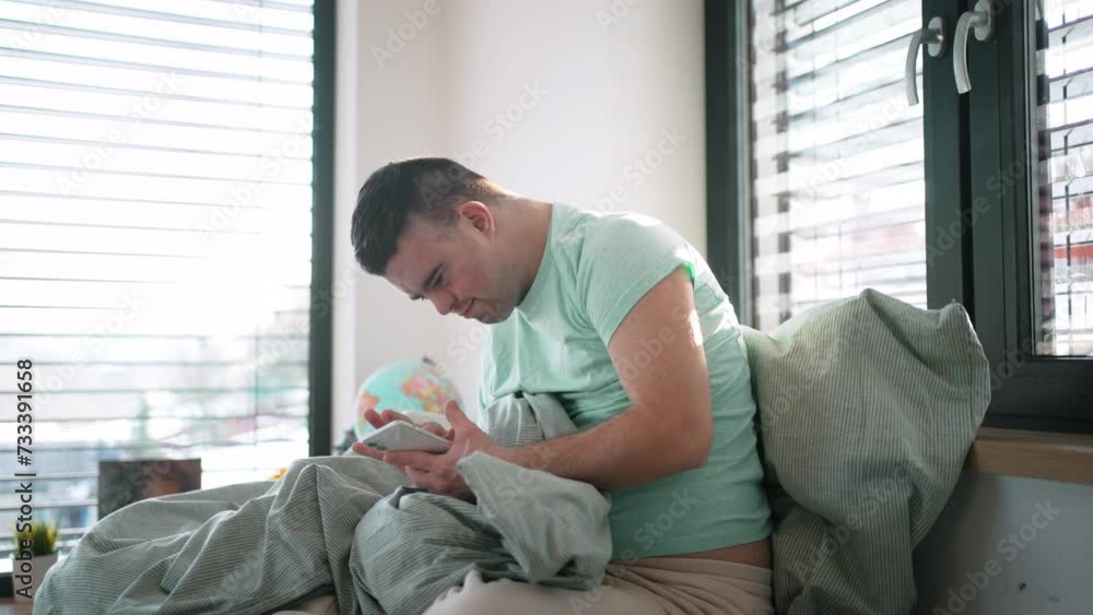 Young man with down syndrome lying in bed, looking at smartphone in morning. Morning routine for man with disability. Young man with down syndrome lying in bed, looking at smartphone in morning. Morning routine for man with disability.