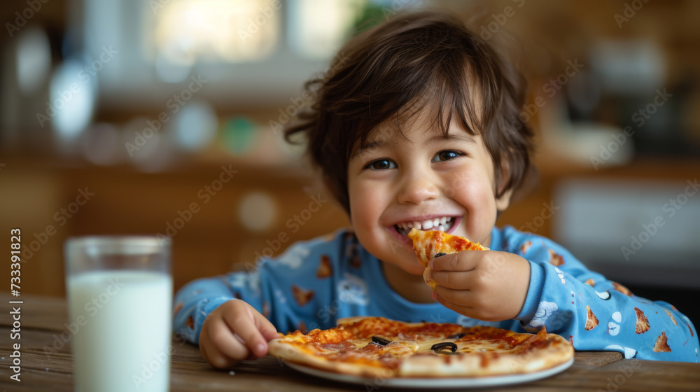 A smiling child in casual clothing is joyfully eating a slice of pizza, with a glass of milk on the table in a home kitchen setting.