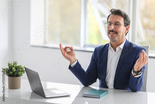 A relaxed businessman in a blue suit practices meditation at his office desk with eyes closed photo