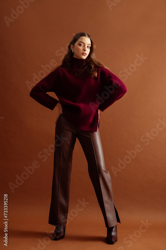 Fashionable confident woman wearing burgundy color knitted sweater, faux leather pants, pointed toe boots, posing on brown background. Full-length studio fashion portrait