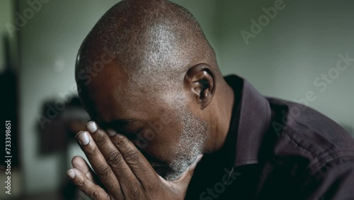 One depressed senior man covers face in shame and regret in moody gloomy bedroom. African American person struggles with poverty and mental illness, close-up face feeling hopeless
