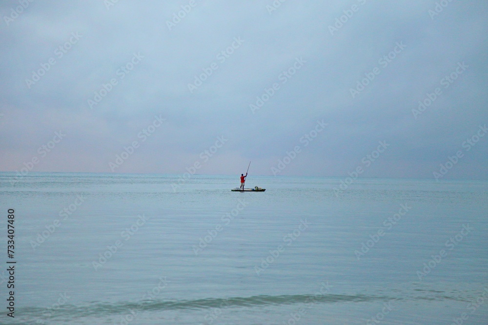 Solitude an fisherman alone over a raft in  wide blue sea.  Emptyness in early day, foggy sky mixing color with sea on horizon line