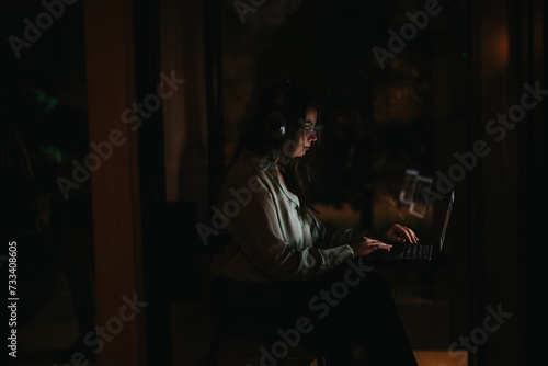 Focused young professional woman using a laptop in a dimly lit office space, working beyond office hours.