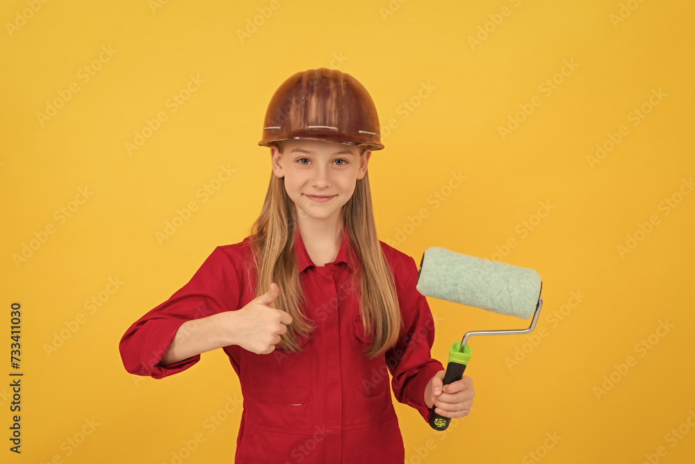 teen child smile in builder hard hat with paint roller on yellow wall. thumb up teen child smile in builder hard hat with paint roller on yellow wall. thumb up