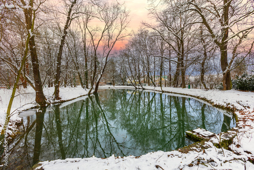 A snowy winter landscape with a small pond and tall trees growing on the shore. Reflections of trees and sky on the surface of the pond.