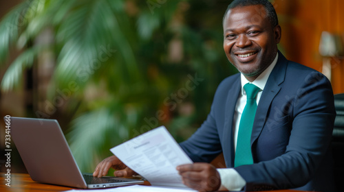 A smiling businessman in a suit is sitting at an office desk, holding a document with a laptop open in front of him.