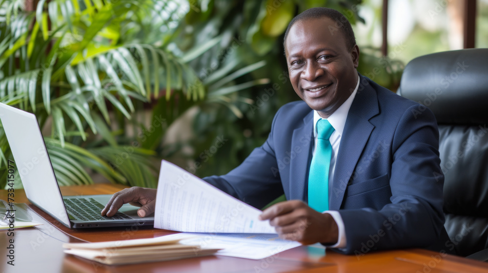 A smiling businessman in a suit is sitting at an office desk, holding a document with a laptop open in front of him. A smiling businessman in a suit is sitting at an office desk, holding a document with a laptop open in front of him.