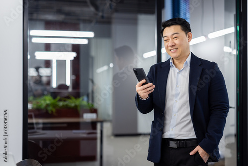 Asian businessman standing inside office with phone in hands, boss in business suit typing text message and reading social media, mature man using app on smartphone.