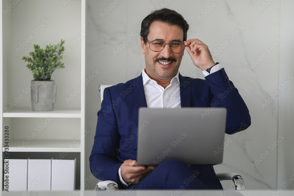 Confident and cheerful mature businessman in blue suit adjusting eyeglasses while working Confident and cheerful mature businessman in blue suit adjusting eyeglasses while working