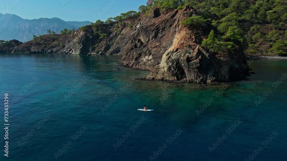 Female paddleboarder moving on sup paddle board calmly through blue waters below cliffs in Mediterranean sea. Aerial wide shot Female paddleboarder moving on sup paddle board calmly through blue waters below cliffs in Mediterranean sea. Aerial wide shot