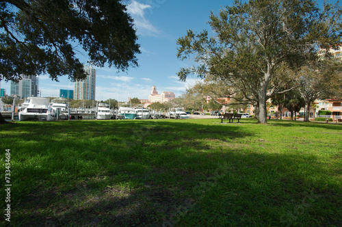 Low angle horizontal view over green grass at Vinoy Park in St. Petersburg, FL. Trees in foreground and sides with boats in water and buildings with blue and white sky later afternoon sun. © Del Harper