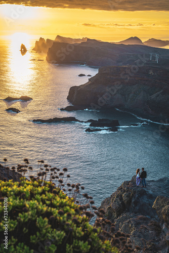 Tourist couple enjoys sunrise from vantage point on steep cliff above seascape and along rugged foothills of Madeira's coastline with wind turbines. Ponta do Bode, Madeira Island, Portugal, Europe.