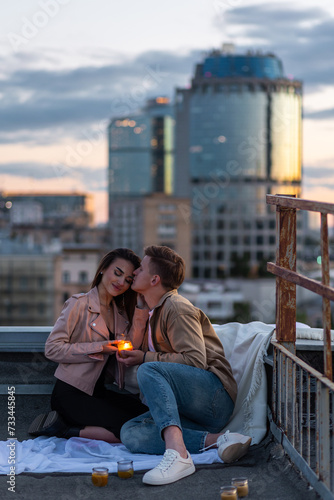 Surprise date on rooftop with urban cityscape and skyscrapers on background. Happy young loving couple drinking wine having romantic candlelit dinner celebrating anniversary or Valentines Day